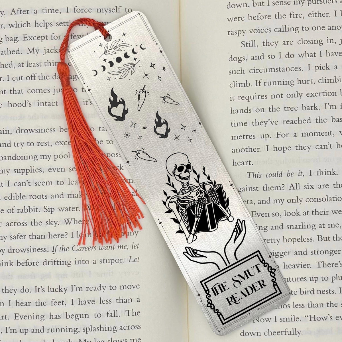 A brushed silver The Smut Reader Metal Bookmark on and open book with a red tassel
