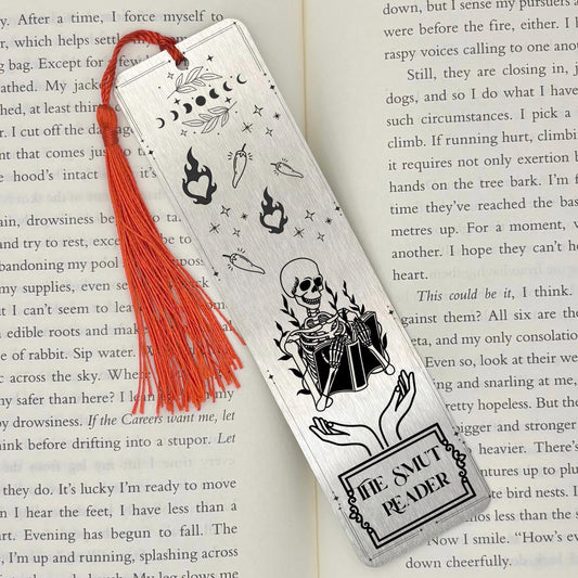 A brushed silver The Smut Reader Metal Bookmark on and open book with a red tassel