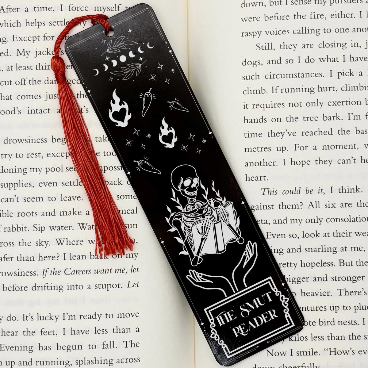 A black The Smut Reader Metal Bookmark on and open book with a red tassel