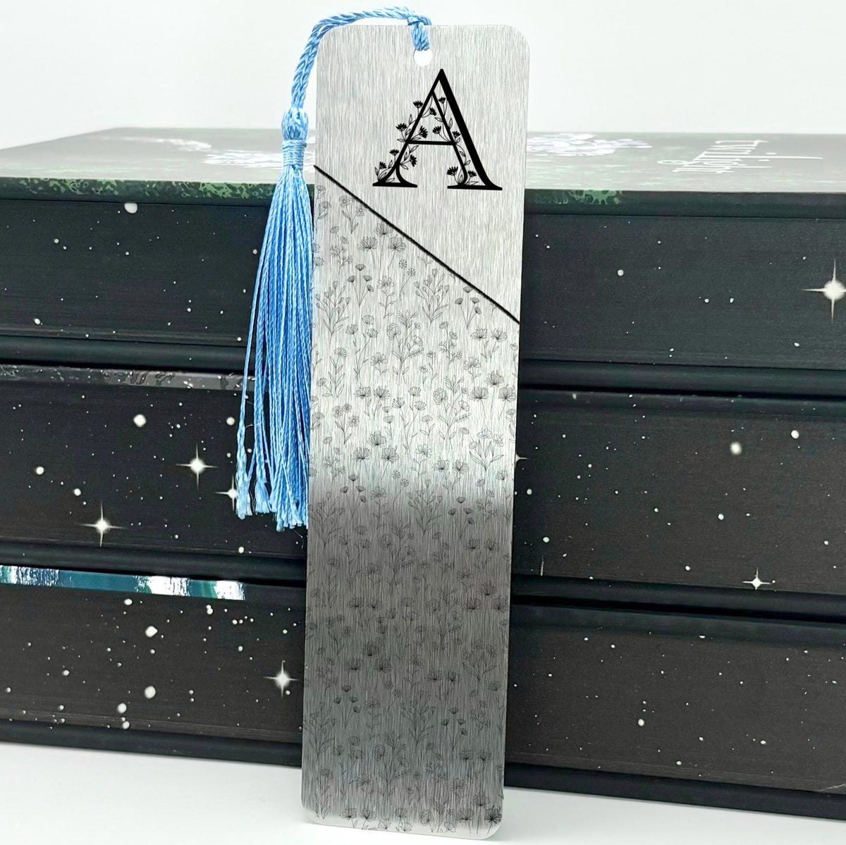 A brushed silver Personalised Floral Initial Metal Bookmark with light blue tassel standing against stack of books