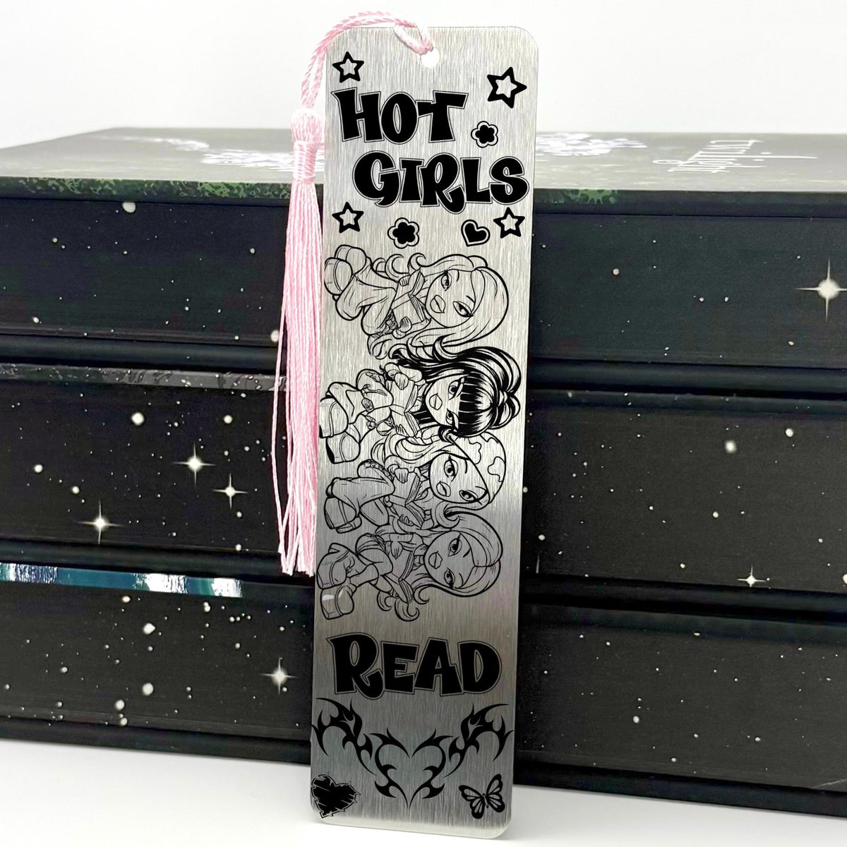 A brushed silver Y2K Hot Girls Read Metal Bookmark with pink tassel standing against a backdrop of books