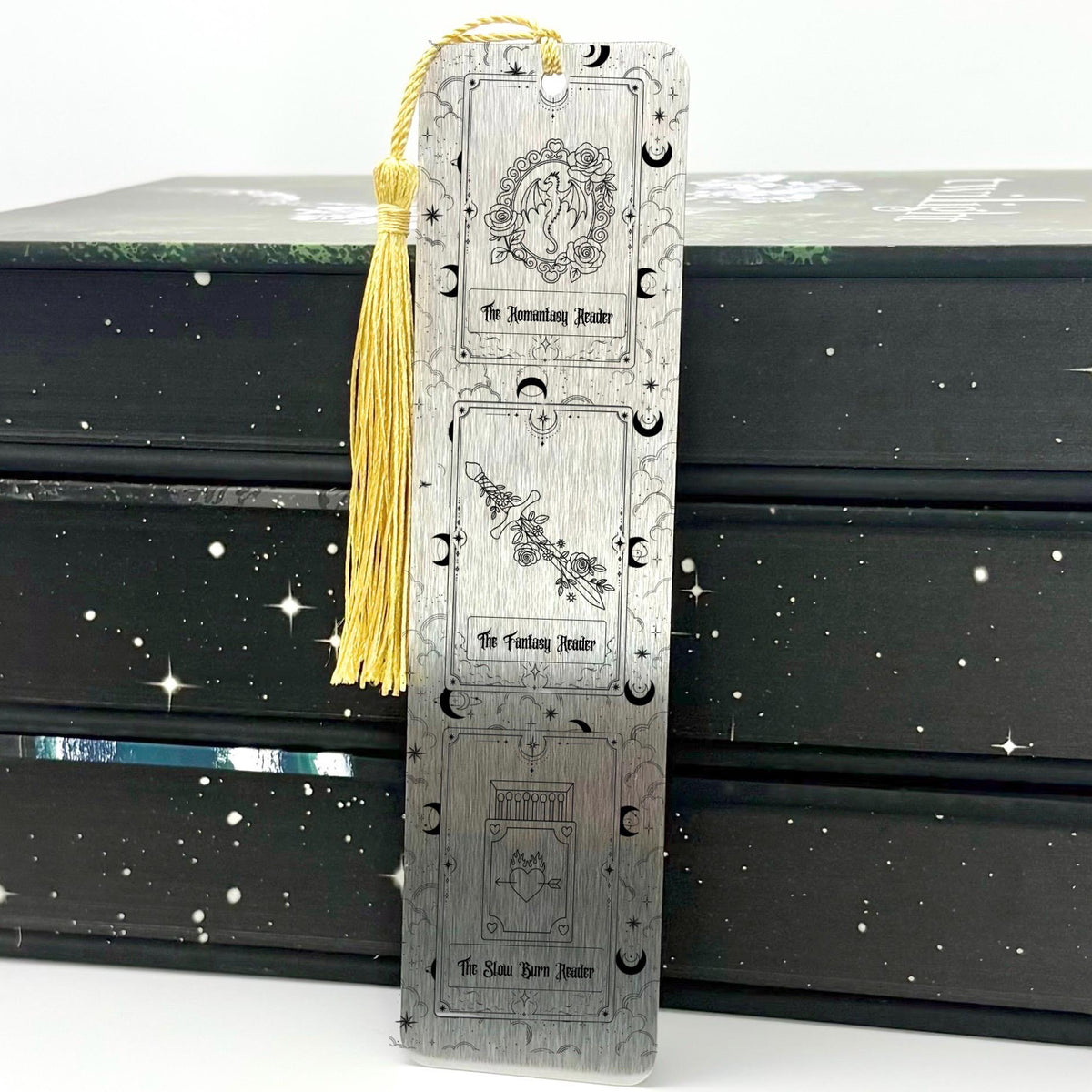 A brushed silver Fantasy Romantasy Slow Burn Metal Bookmark with gold tassel standing against a backdrop of books