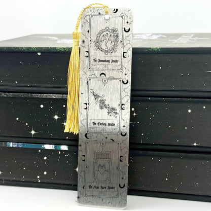 A brushed silver Fantasy Romantasy Slow Burn Metal Bookmark with gold tassel standing against a backdrop of books