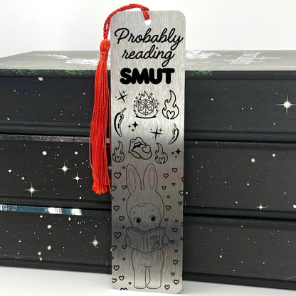 A brushed silver Probably Reading Smut Metal Bookmark with red tassel standing against a stack of books