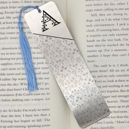 A brushed silver Personalised Floral Initial Metal Bookmark on an open book with light blue tassel