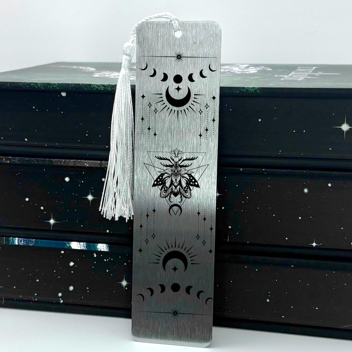 A brushed silver Celestial Moon and Stars Metal Bookmark with white tassel standing against a backdrop of books